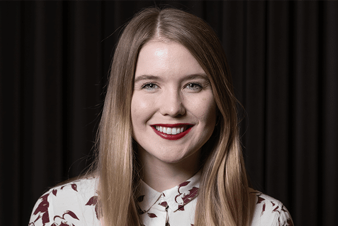 A person with long blonde hair is smiling at the camera, as if preparing for a hiring assessment. They are wearing a white shirt with a maroon leaf pattern. The background is dark with vertical lines, adding an air of mystery to the scene.