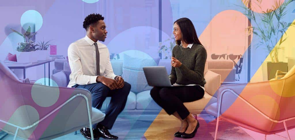 A man in a white shirt and a woman in a green sweater are sitting and talking in a colorful, modern office space. She is holding a laptop. The chairs are pink and yellow, and abstract shapes overlay the image.