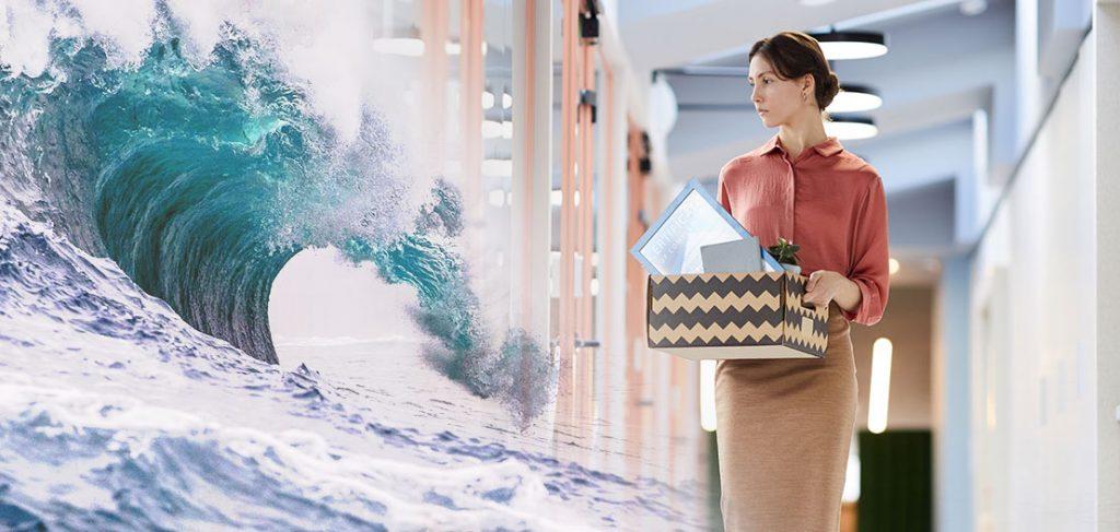 A woman wearing a red blouse and beige skirt holds a box with office items, walking in a hallway. A large ocean wave is digitally overlaid on the left side, blending with the office environment.