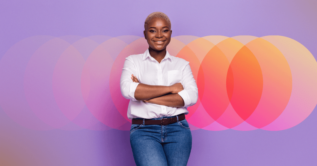 Smiling confidently, a person stands with arms crossed against a vibrant background of overlapping circular patterns in shades of orange, pink, and purple. Dressed in a white shirt and blue jeans, they exude an aura of growth and employee performance development.