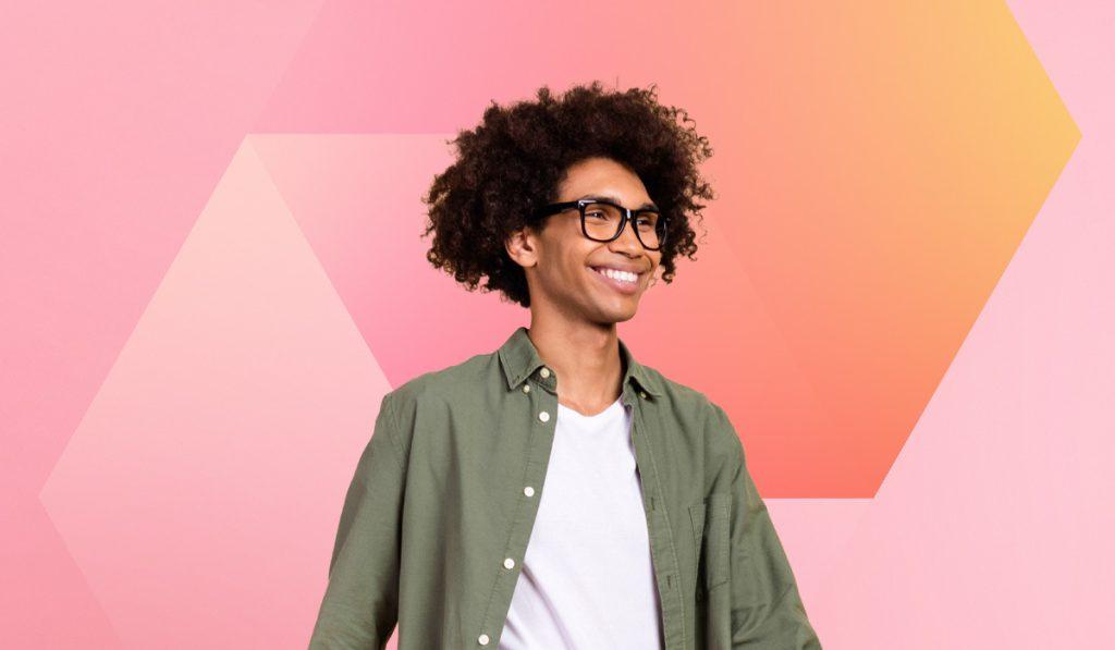 A smiling person with curly hair and glasses, donning a green shirt over a white T-shirt, stands confidently against a pink and orange geometric backdrop—reflecting the vibrant spirit often seen nurtured through employee development programs.