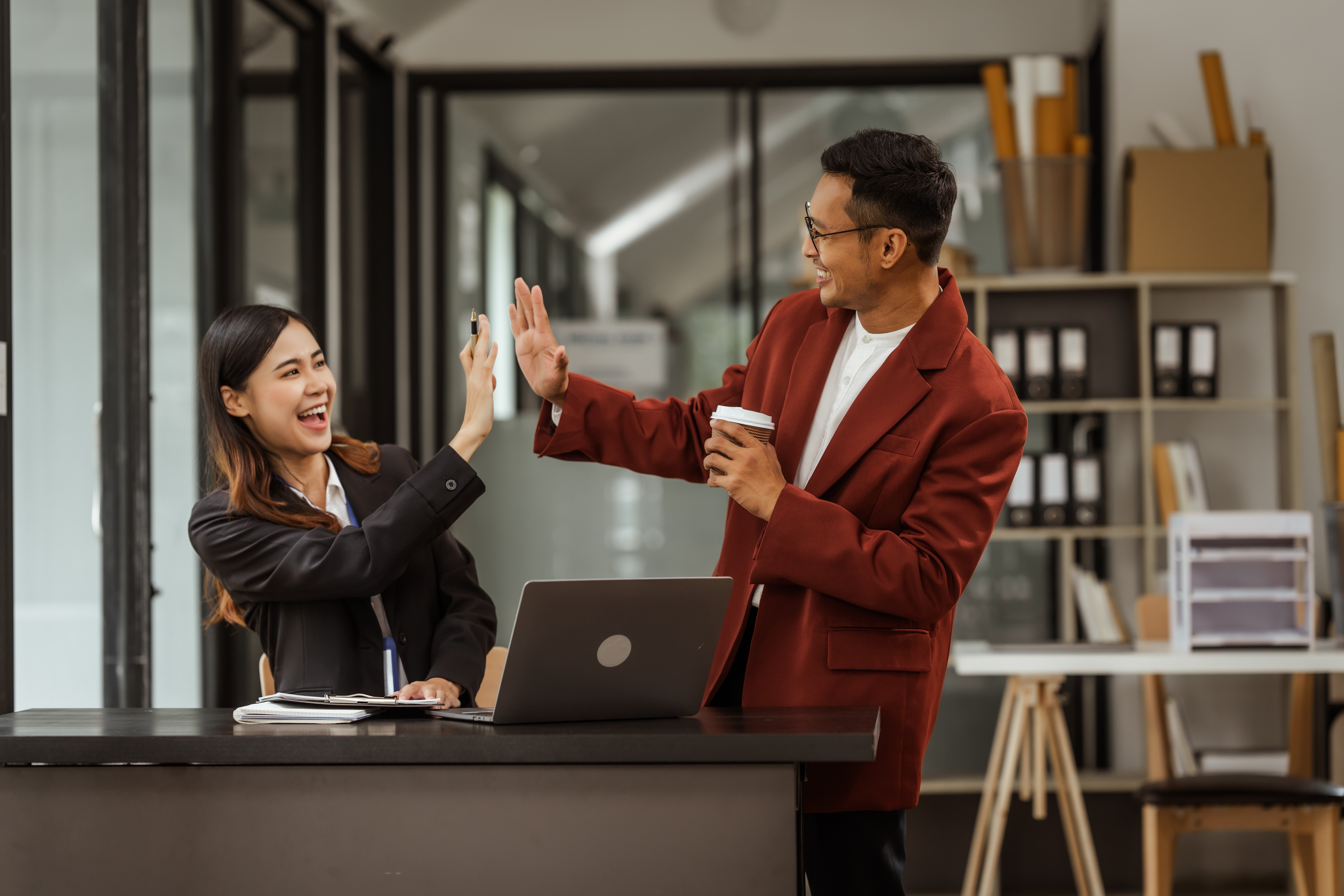 A diverse group of two colleagues, a man and a woman, sharing a joyful high-five in a modern office setting, representing efforts to reduce employee turnover through engagement and a positive workplace culture.