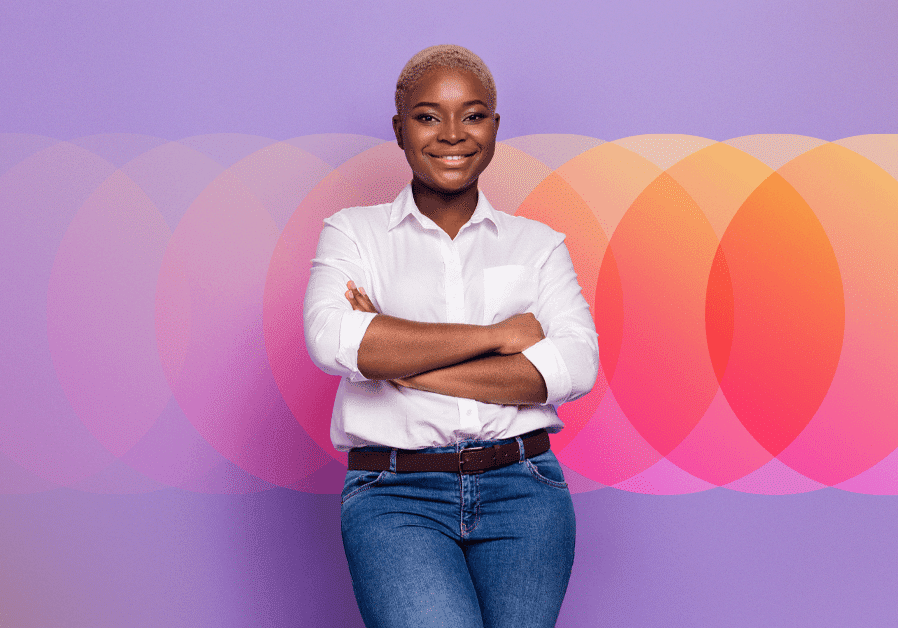 Smiling confidently, a person stands with arms crossed against a vibrant background of overlapping circular patterns in shades of orange, pink, and purple. Dressed in a white shirt and blue jeans, they exude an aura of growth and employee performance development.